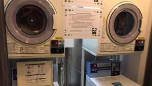 Uma lavanderia com duas máquinas de lavar frontais e uma secadora. Instruções de uso das máquinas estão afixadas na parede.