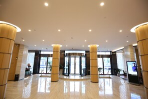 Um saguão de hotel com porta giratória, grandes colunas de madeira, balcão de recepção e uma tela digital exibindo informações.