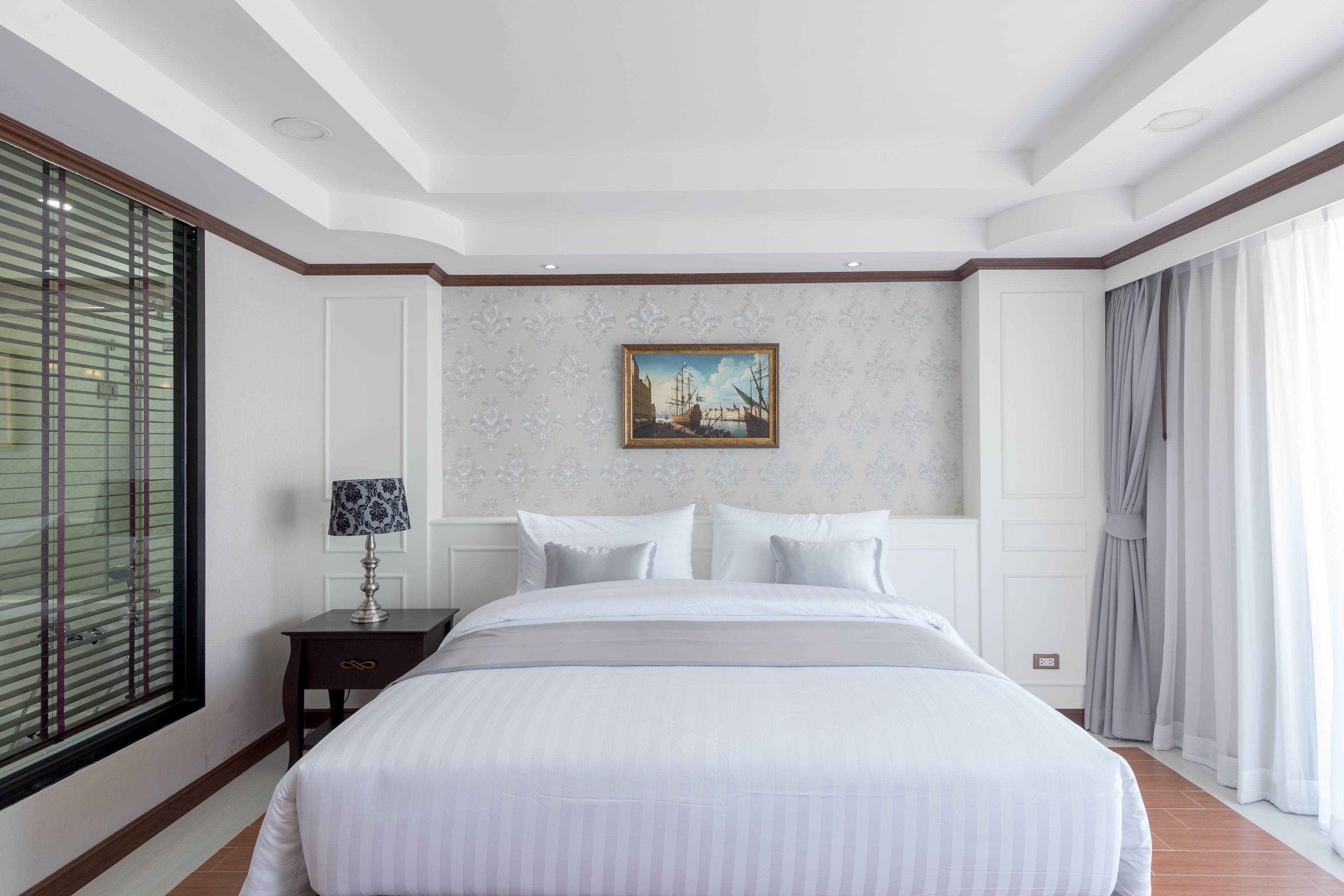 Habitación de hotel ordenada con una cama grande, una mesita de noche pequeña con una lámpara, un cuadro en la pared y una ventana con persianas.