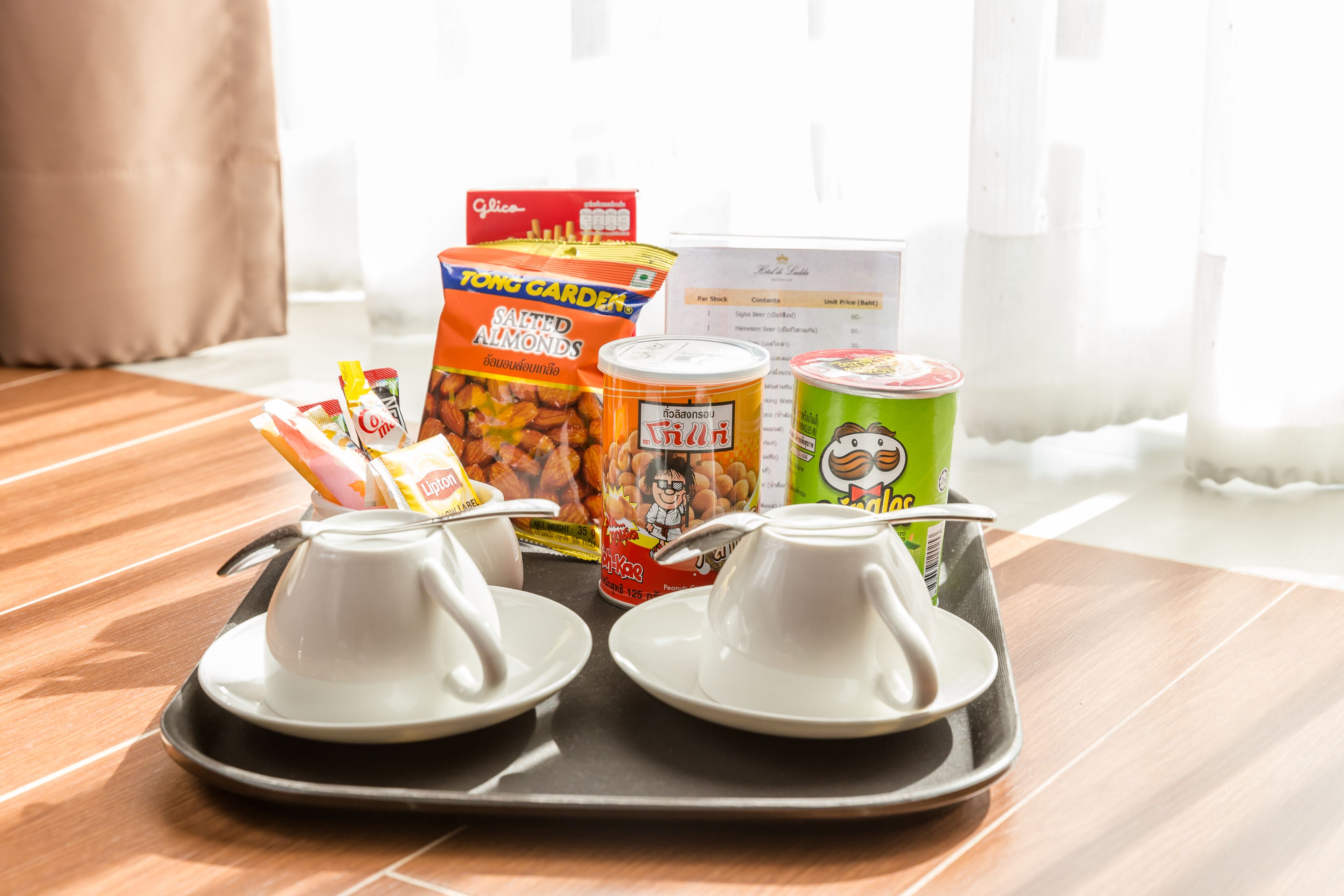A tray with two white cups, a box of salted almonds, a can of fruit, and a green can on a wooden table.