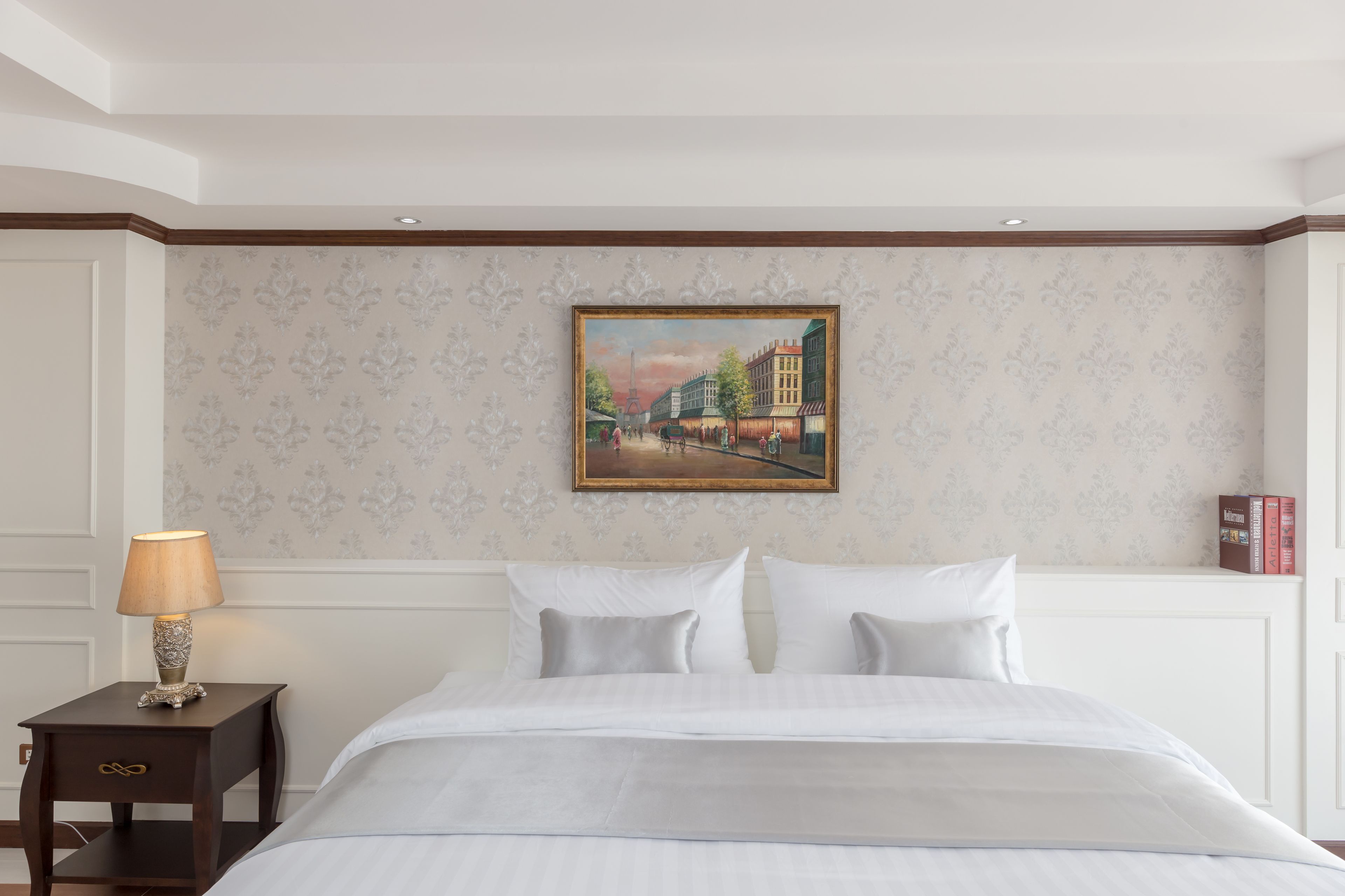 A hotel room with a bed, a nightstand with a lamp, a painting on the wall, and a bookshelf.