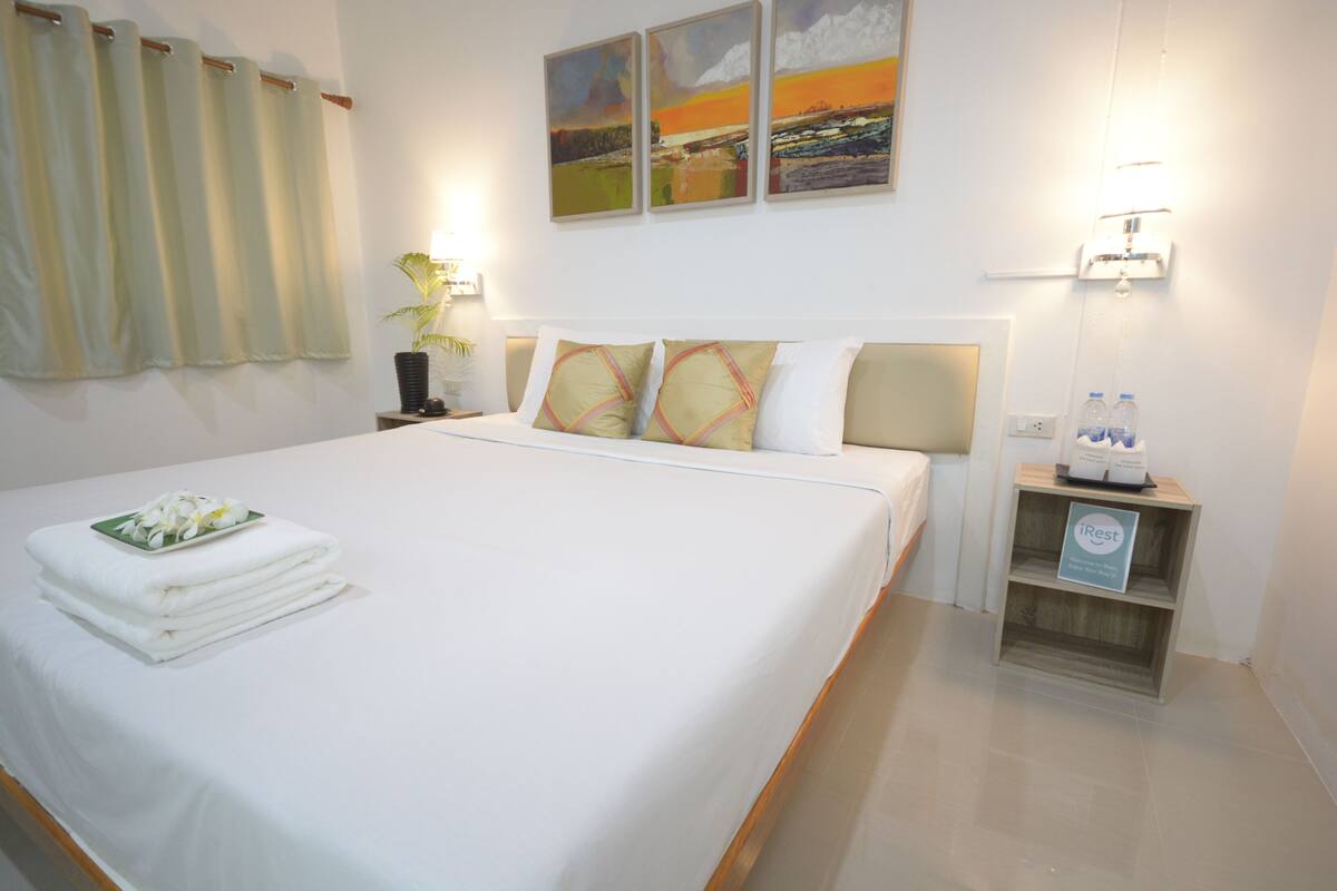 A neatly made bed with white linens and a folded towel, a bedside table with a lamp and a water bottle, and a small shelf with a sign.