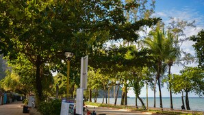 A paved walkway alongside a road with a parked scooter, a signboard, and a view of the sea with palm trees. - iRest Ao Nang Seafront (Krabi)