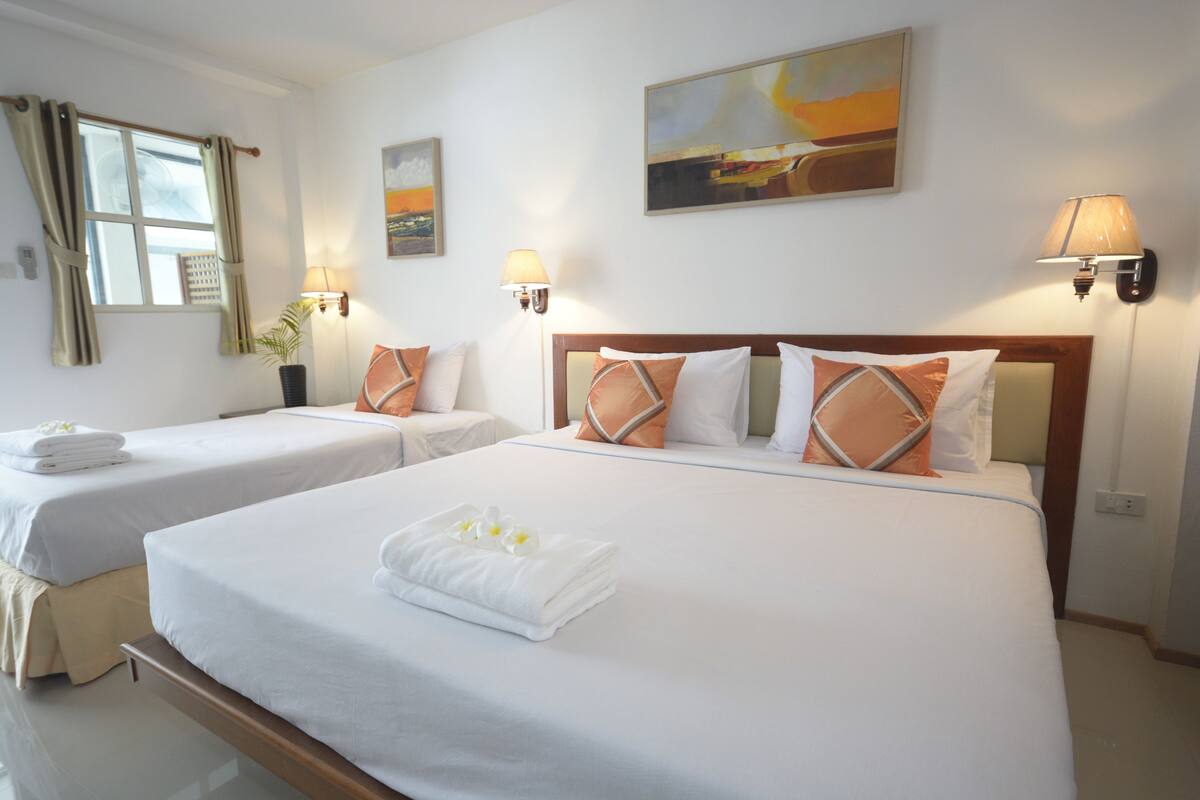 A hotel room with two beds, each with white linens and decorative pillows. There are wall-mounted lamps and a painting above the beds.