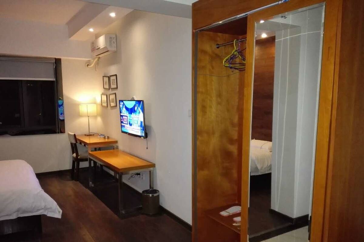 Una habitación de hotel compacta con cama, escritorio, silla, televisión y armario con espejo.