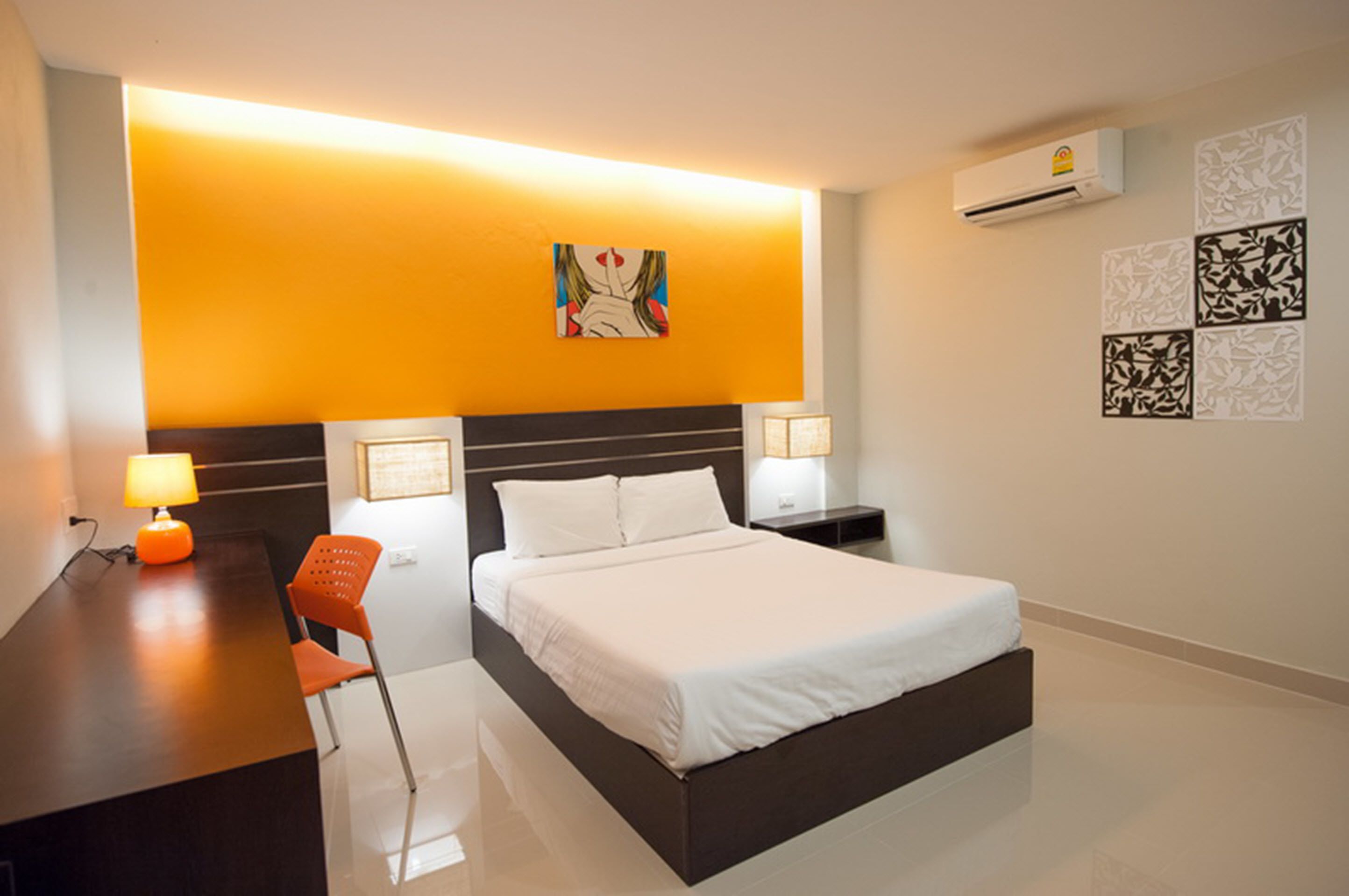 Une chambre d’hôtel comprenant un lit, un bureau, un fauteuil orange, un climatiseur fixé au mur et des œuvres d’art décoratives accrochées au mur.