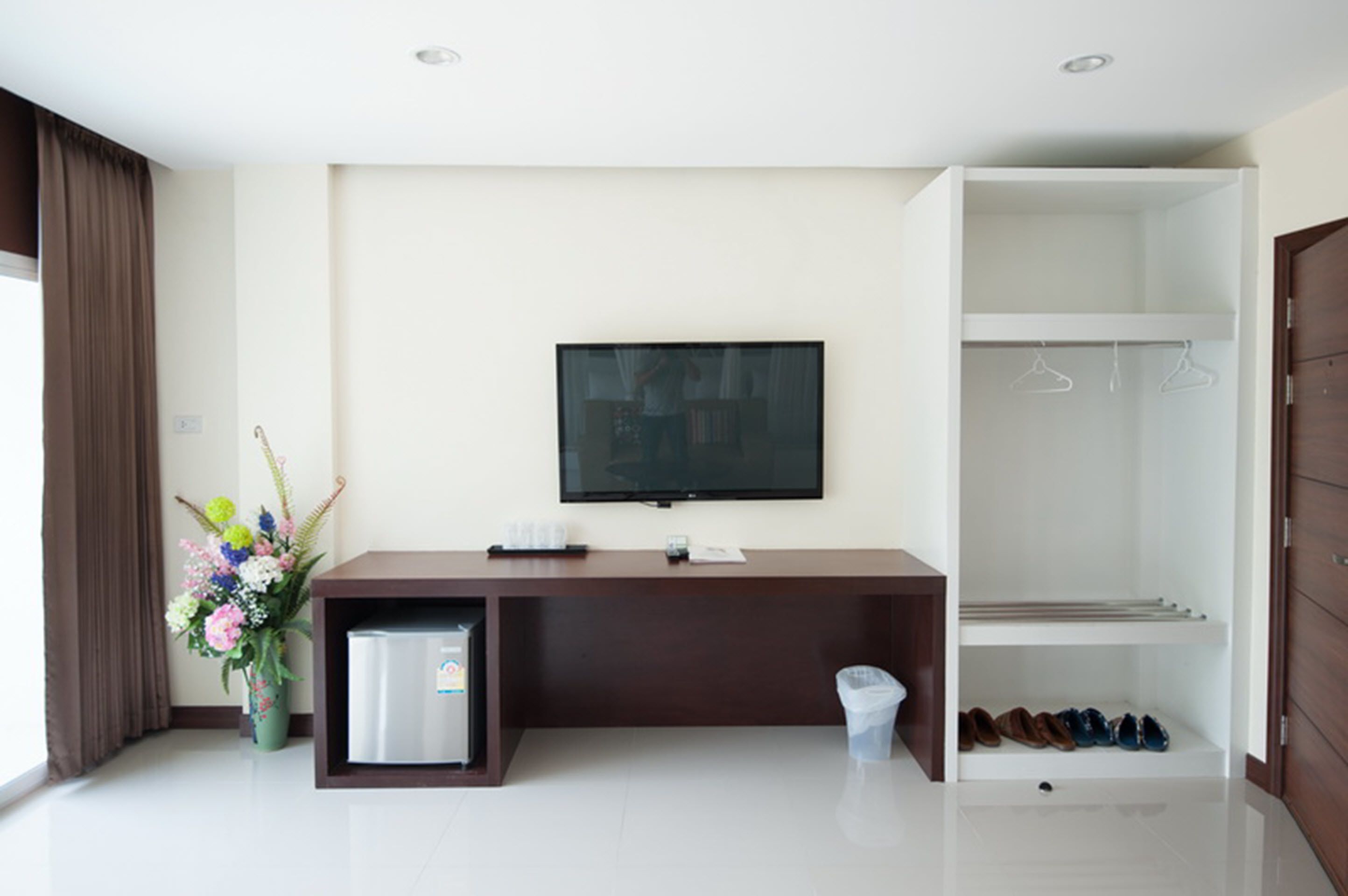 Una habitación de hotel moderna con televisión de pantalla plana montada en la pared, un refrigerador pequeño, un escritorio con un cesto de basura, un perchero para zapatos y un ventanal con cortinas.