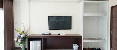 Una habitación de hotel moderna con televisión de pantalla plana montada en la pared, un refrigerador pequeño, un escritorio con un cesto de basura, un perchero para zapatos y un ventanal con cortinas.