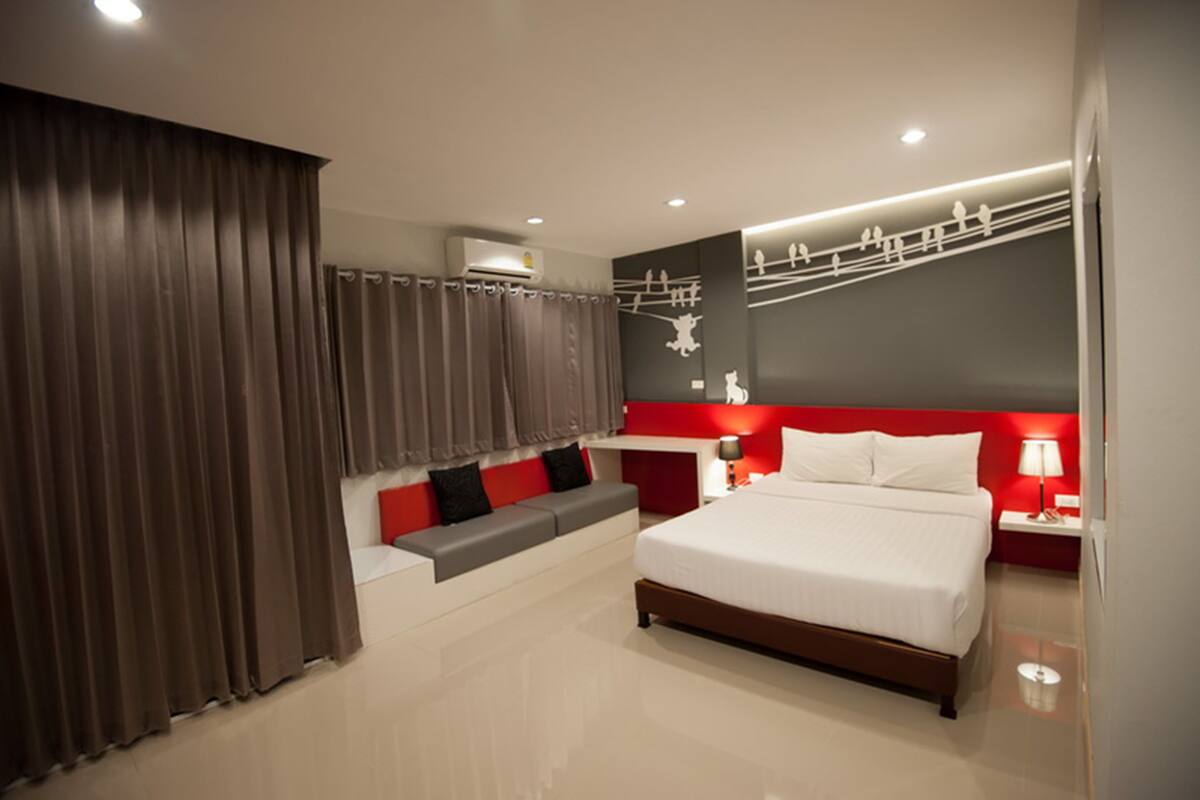 A modern hotel room with a large bed, a seating area with cushions, a red accent wall, and an air conditioning unit.