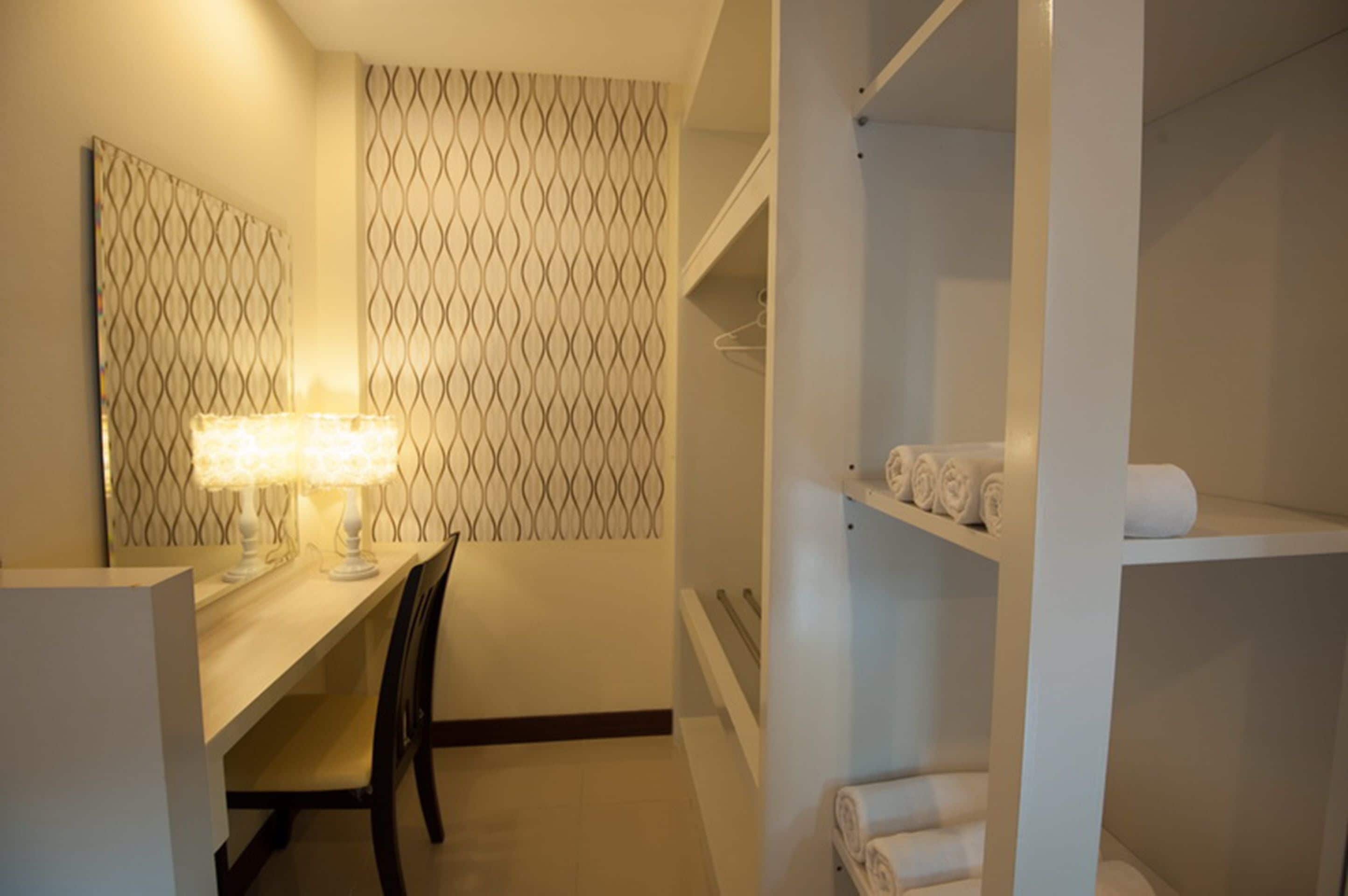 A hotel room with a desk, chair, and a wall-mounted vanity with a mirror. There are shelves with towels and a patterned wall design.