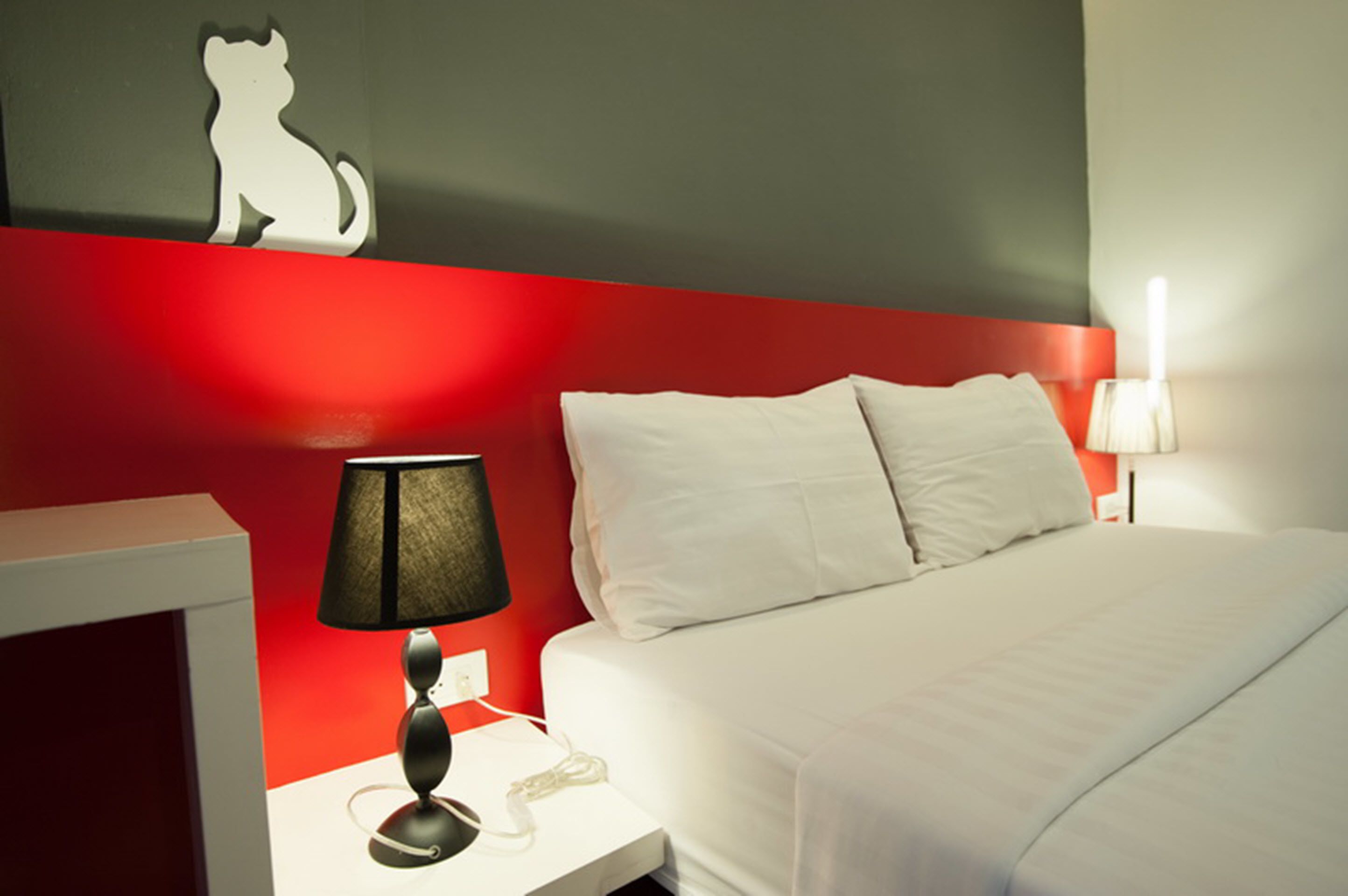Habitación de hotel con una pared de acento roja, una cama blanca, una lámpara negra y una mesita de noche blanca con un teléfono.
