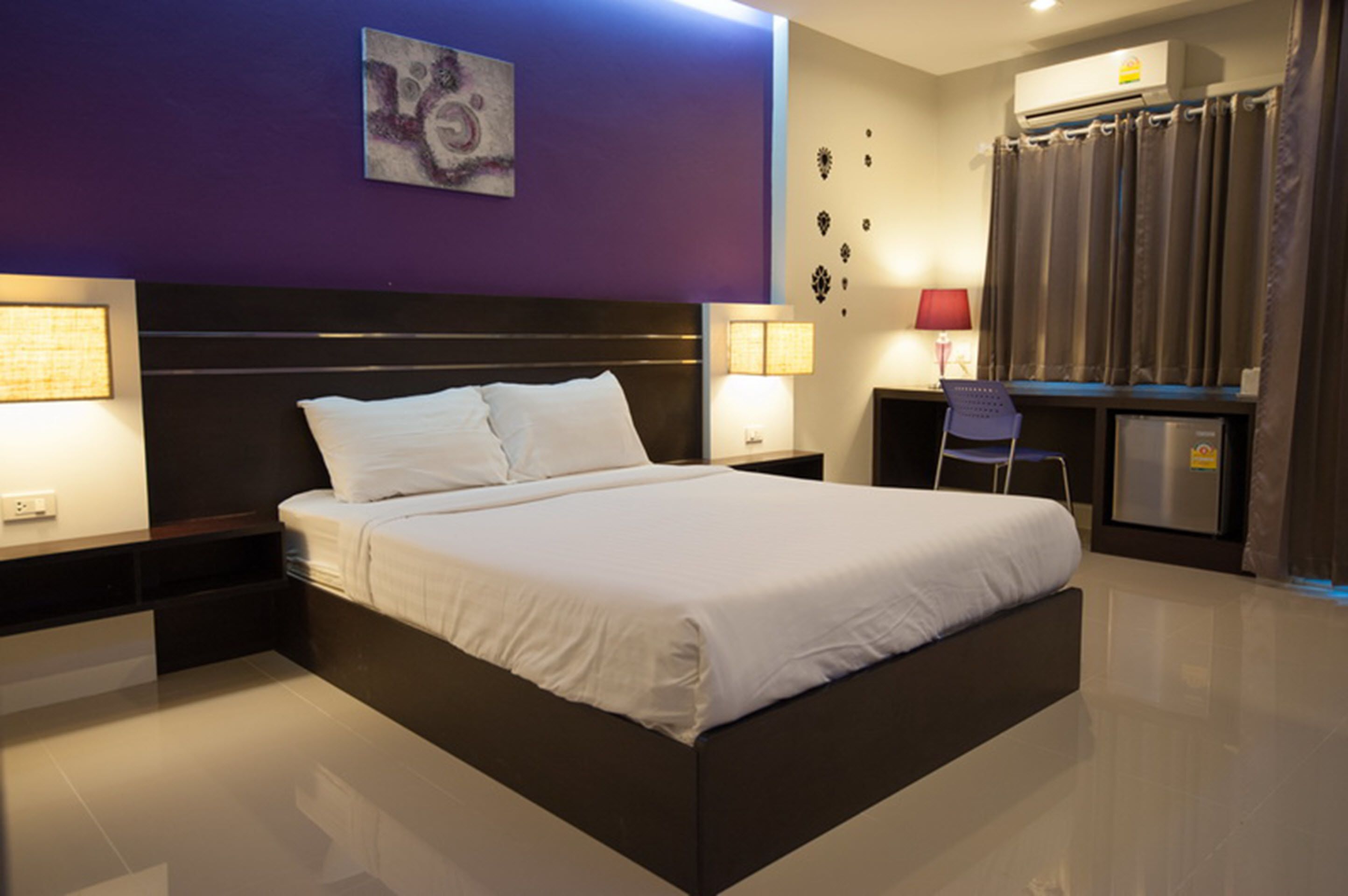 A modern hotel room with a large bed, bedside lamps, a desk with a chair, and an air conditioning unit.