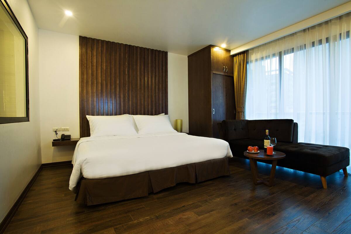 Habitación de hotel con una cama grande, un sillón oscuro, una mesita con una botella y un vaso, y una ventana con cortinas traslúcidas.