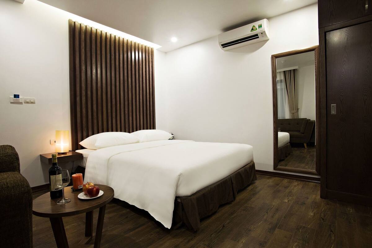 A hotel room with a bed, bedside table with a bottle, glass, and fruit, a wall-mounted air conditioner, and a mirror.