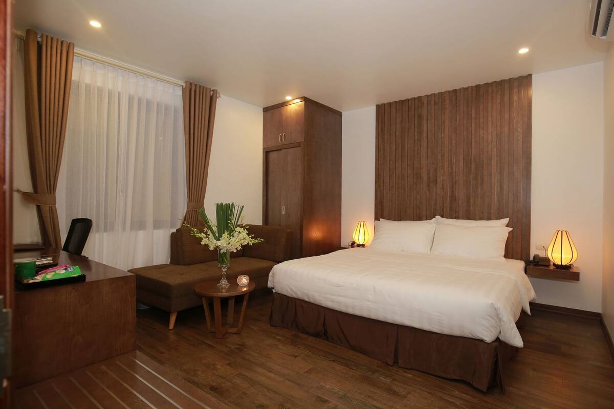 A hotel room with a bed, bedside tables, a desk, a chair, a small table with a vase of flowers, and a wooden headboard.