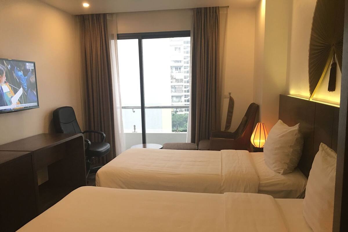 A hotel room with two beds, a desk, a chair, a TV, and a window with a view of buildings.