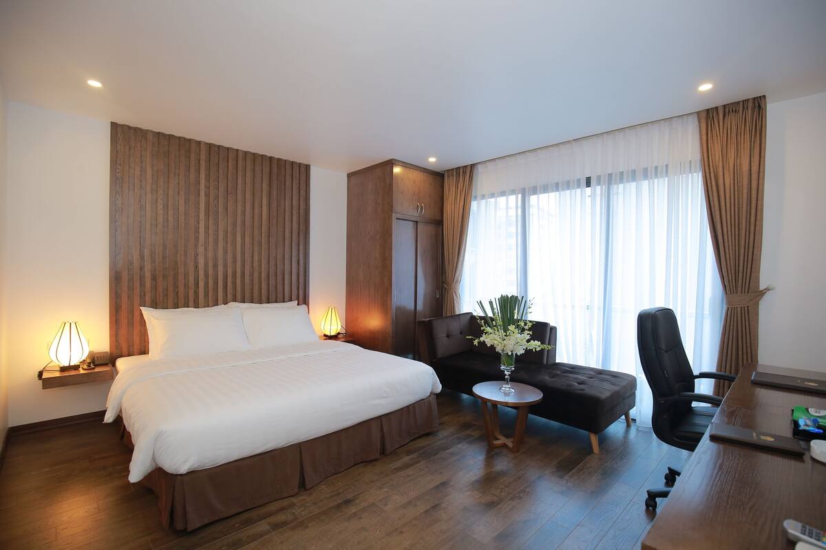 Habitación de hotel con una cama grande, un escritorio, una silla, una mesita con un florero y una ventana con cortinas.
