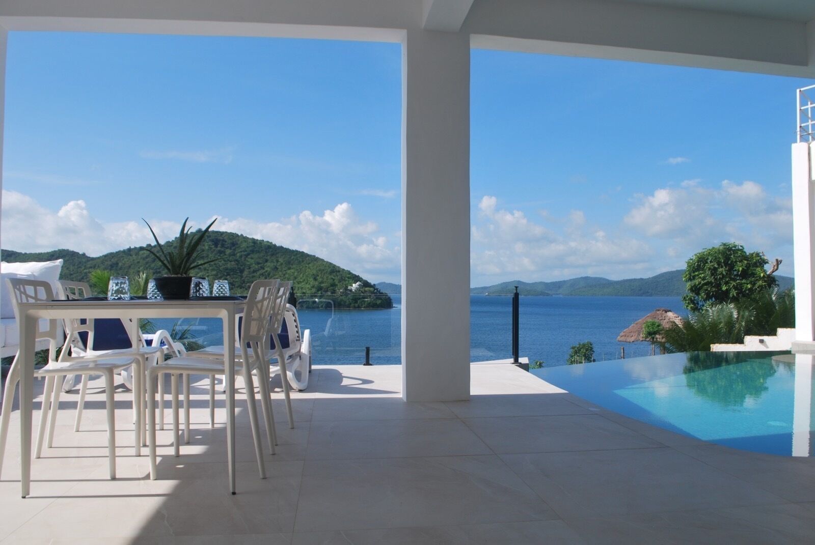 A terrace with a table and chairs overlooking a pool and a scenic view of a body of water and a green island.