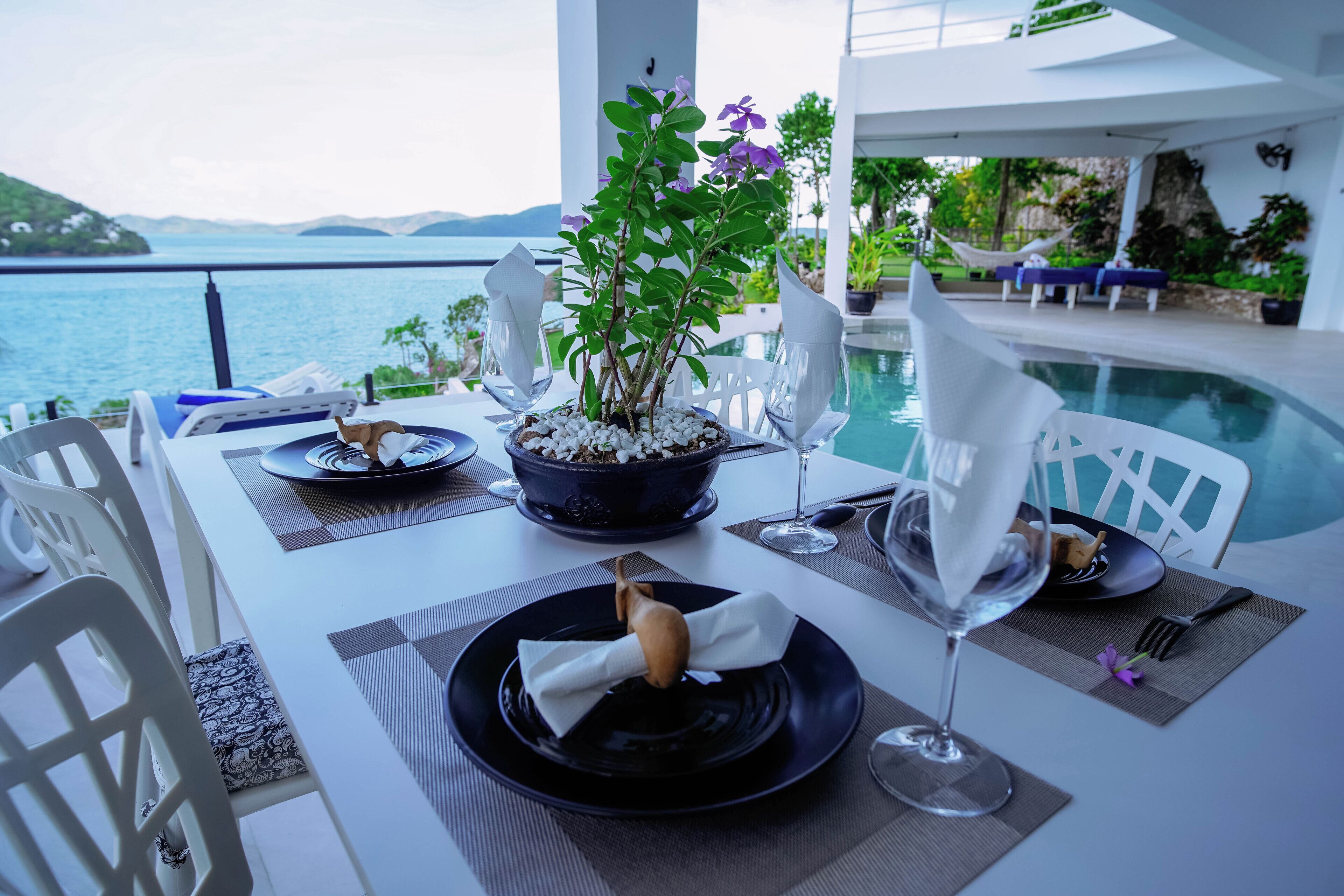 A table set for a meal with a view of the sea, a plant centerpiece, and a dining area with white chairs and a pool in the background.