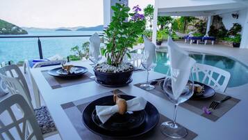 A table set for a meal with a view of the sea, a plant centerpiece, and a dining area with white chairs and a pool in the background.
