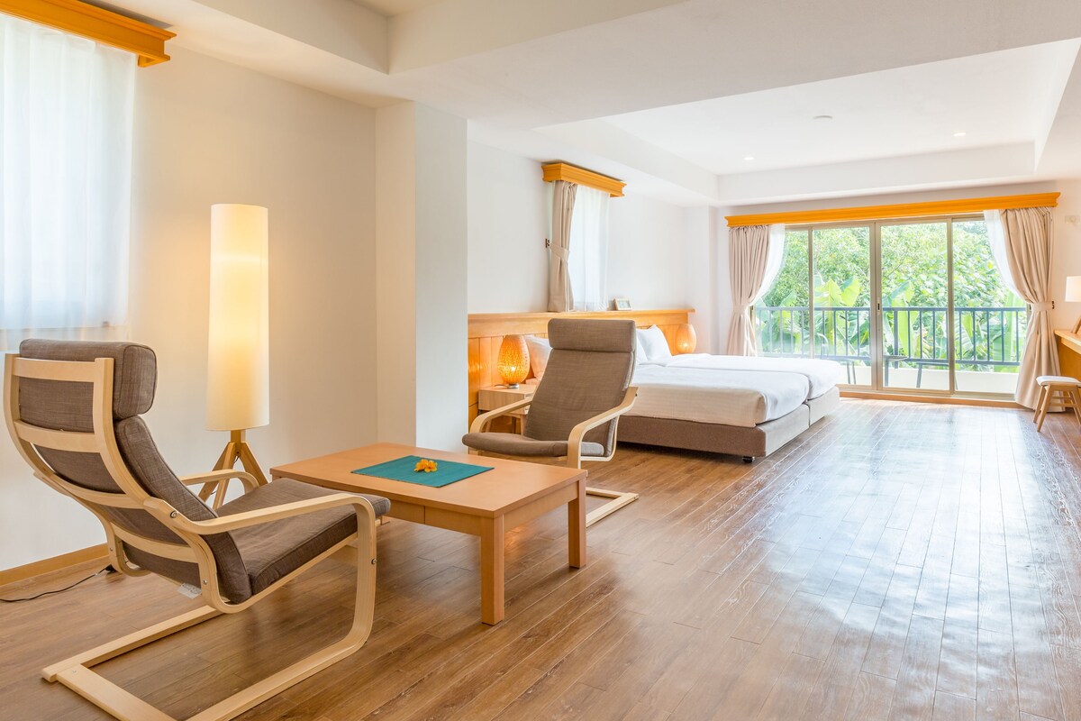 Habitación de hotel con cama, dos sillones, una mesa y un gran ventanal con cortinas.