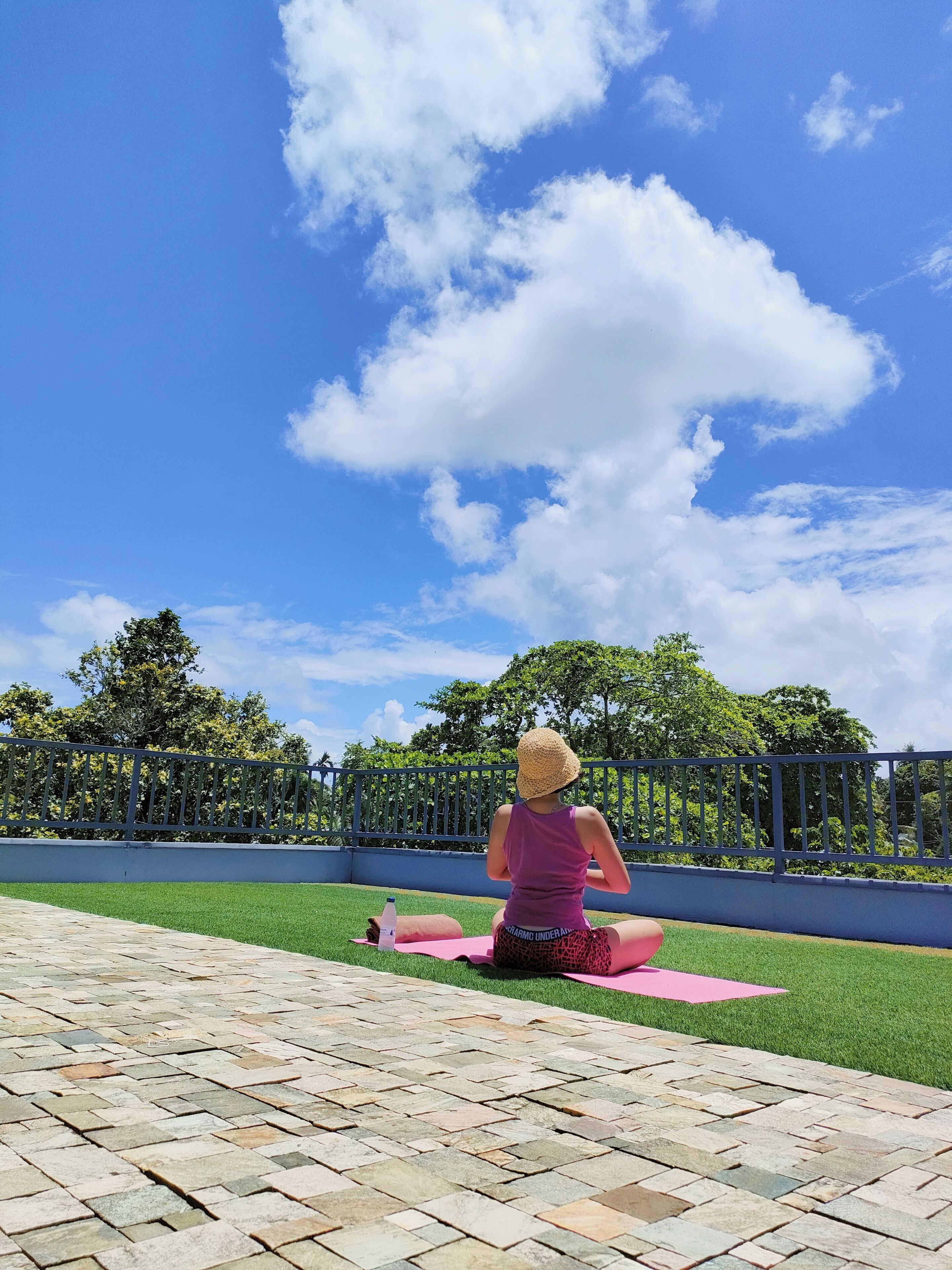 A person sitting on a pink mat outdoors with a clear blue sky and trees in the background.