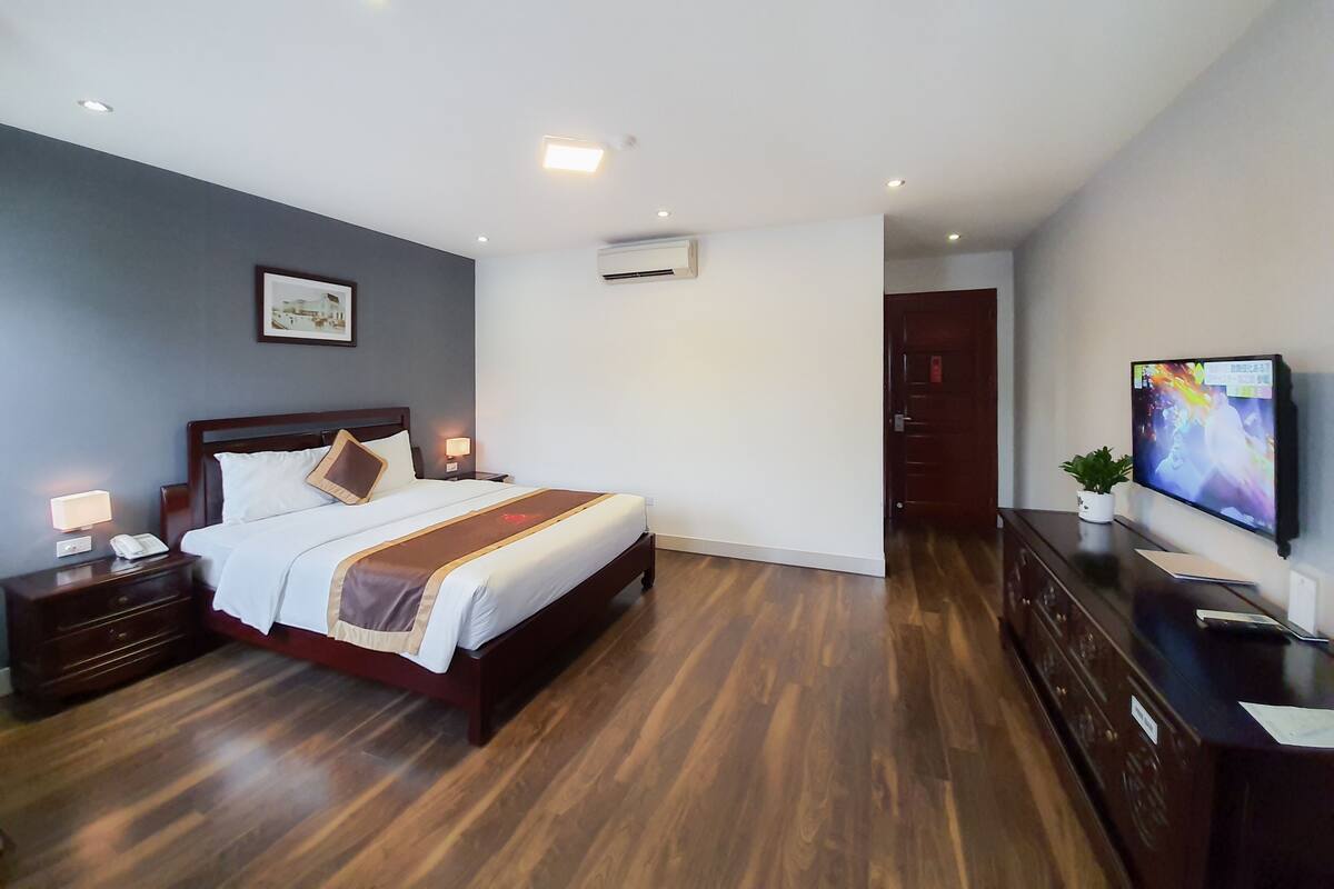 Una habitación de hotel con una cama de madera, una mesita de noche con lámpara, un televisor en la pared y un equipo de aire acondicionado.