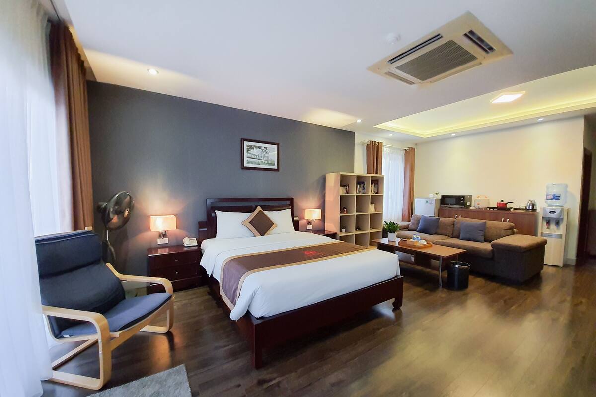 Habitación de hotel con cama, sofá, escritorio, estantería, ventilador y dispensador de agua.