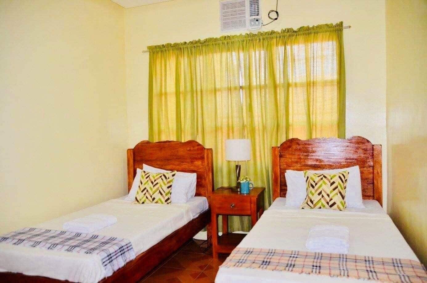 Quarto com duas camas de solteiro, cada uma com cabeceira de madeira, mesa de cabeceira com abajur, e janela com cortinas verdes.