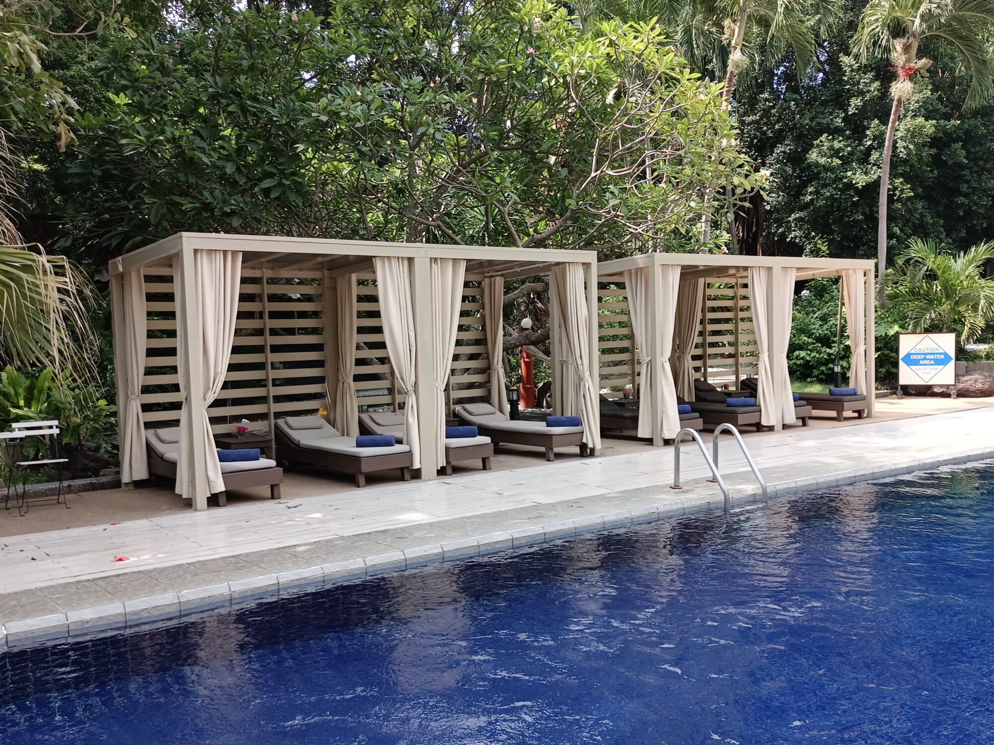 Un espace aménagé au bord de la piscine, comprenant trois chaises longues à l’ombre et un panneau indiquant une « zone à grande profondeur ».