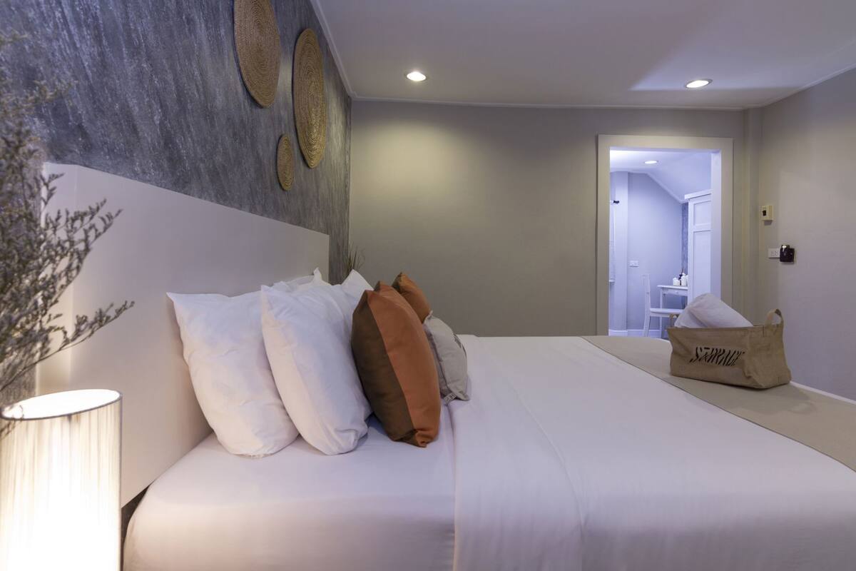 Un dormitorio con cama, almohadas, una lámpara, un adorno de pared tejido y un bolso en el suelo.