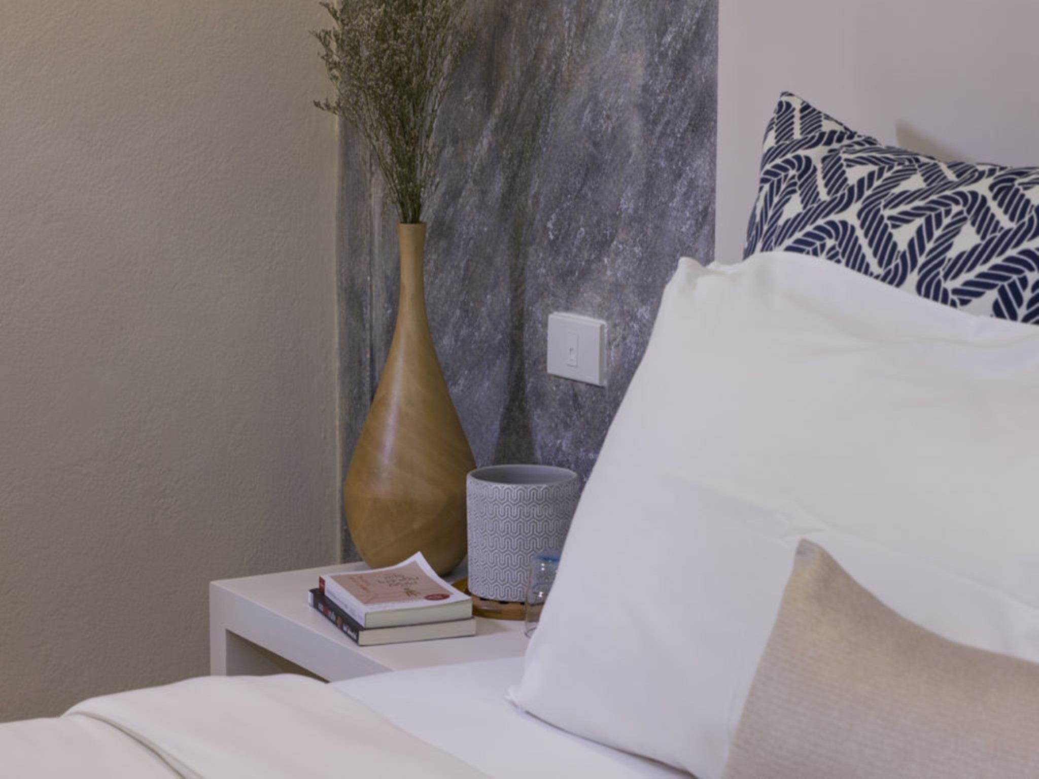 Una cama bien hecha con un cojín blanco y uno estampado, una mesita de noche con un libro y un florero con flores secas, y una pared con textura al fondo.