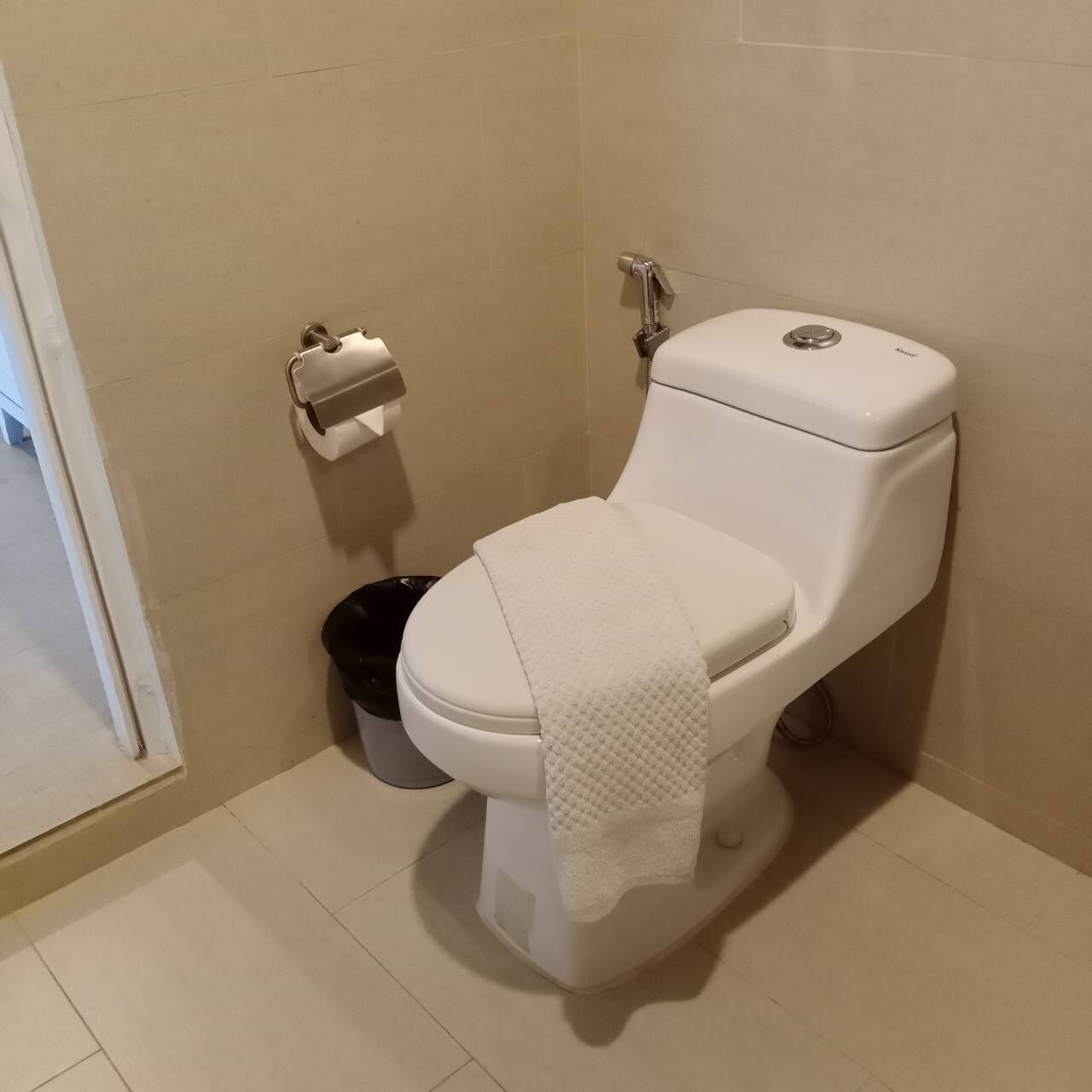 A white toilet with a paper towel holder and a black trash bin in a bathroom with beige tiles.