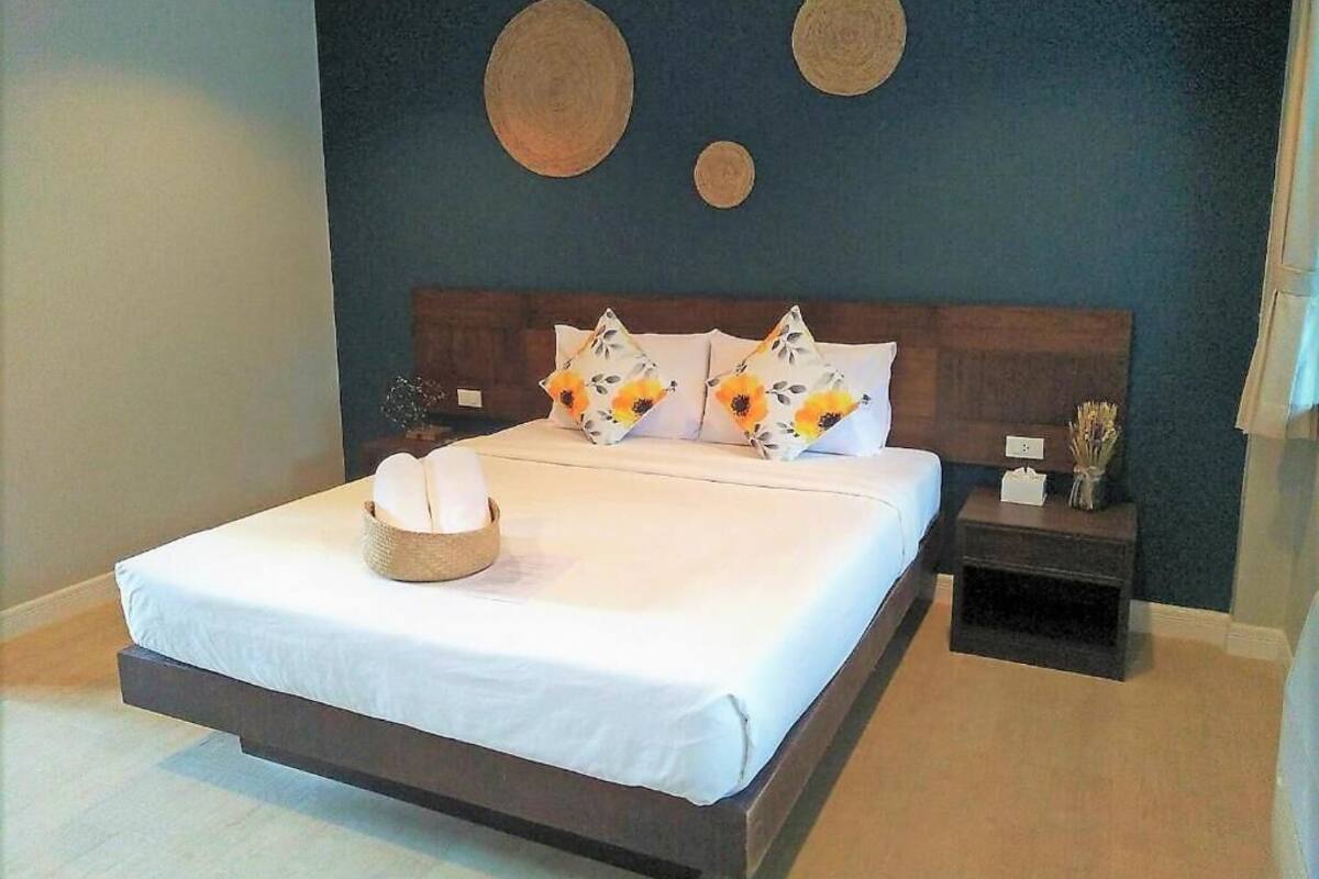 Una cama bien hecha con ropa blanca y almohadillas decorativas, una mesita de noche con un florero y una pared adornada con un arte circular.