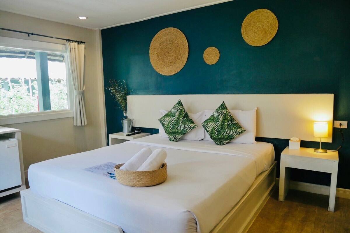 Una habitación de hotel con una cama, una mesita de noche con lámpara, una pequeña mesa con un jarrón y una ventana con cortinas.