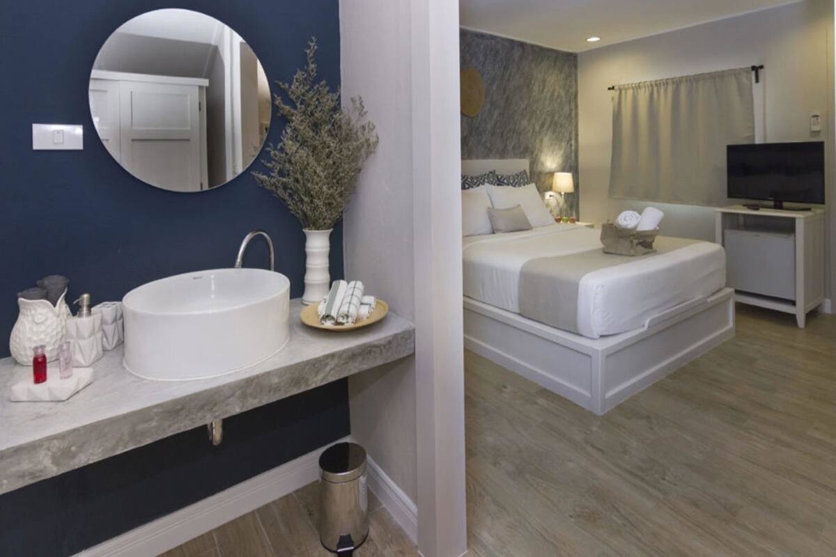 Habitación de hotel con un espejo grande y redondo, un lavamanos blanco, una cama con dos almohadas, una mesita de noche con una lámpara y un televisor.