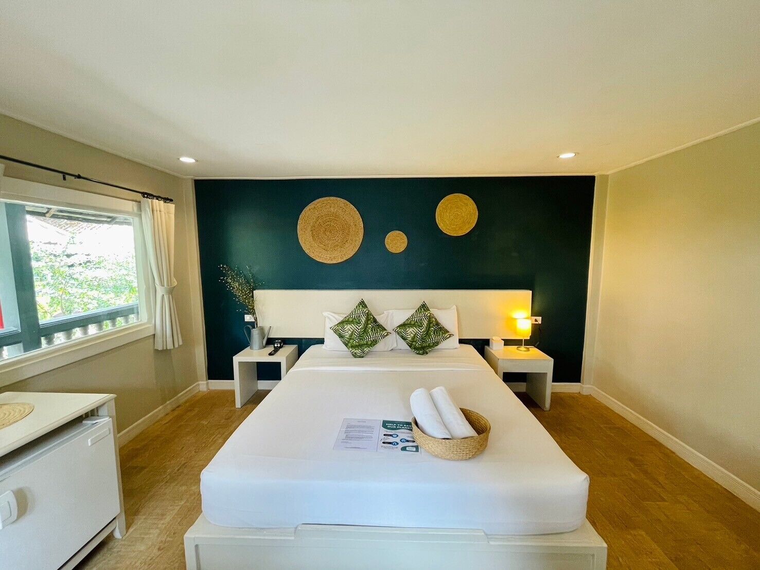 A modern hotel room with a bed, bedside tables, a nightstand, a window with curtains, and a view of greenery outside.