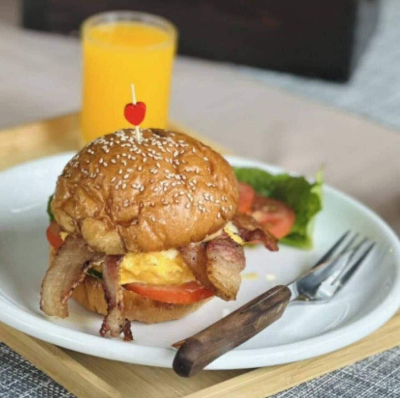En morgenmadstallerken med burger, bacon, æg og tomat, ledsaget af et glas appelsinjuice.