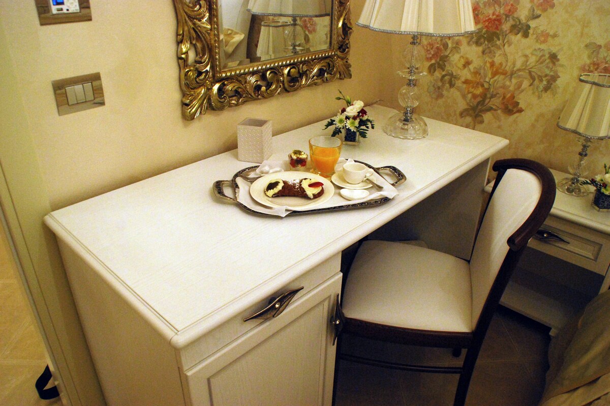 Habitación de hotel con escritorio, silla, espejo y una pequeña mesa con una bandeja de comida, un vaso de jugo y una taza.