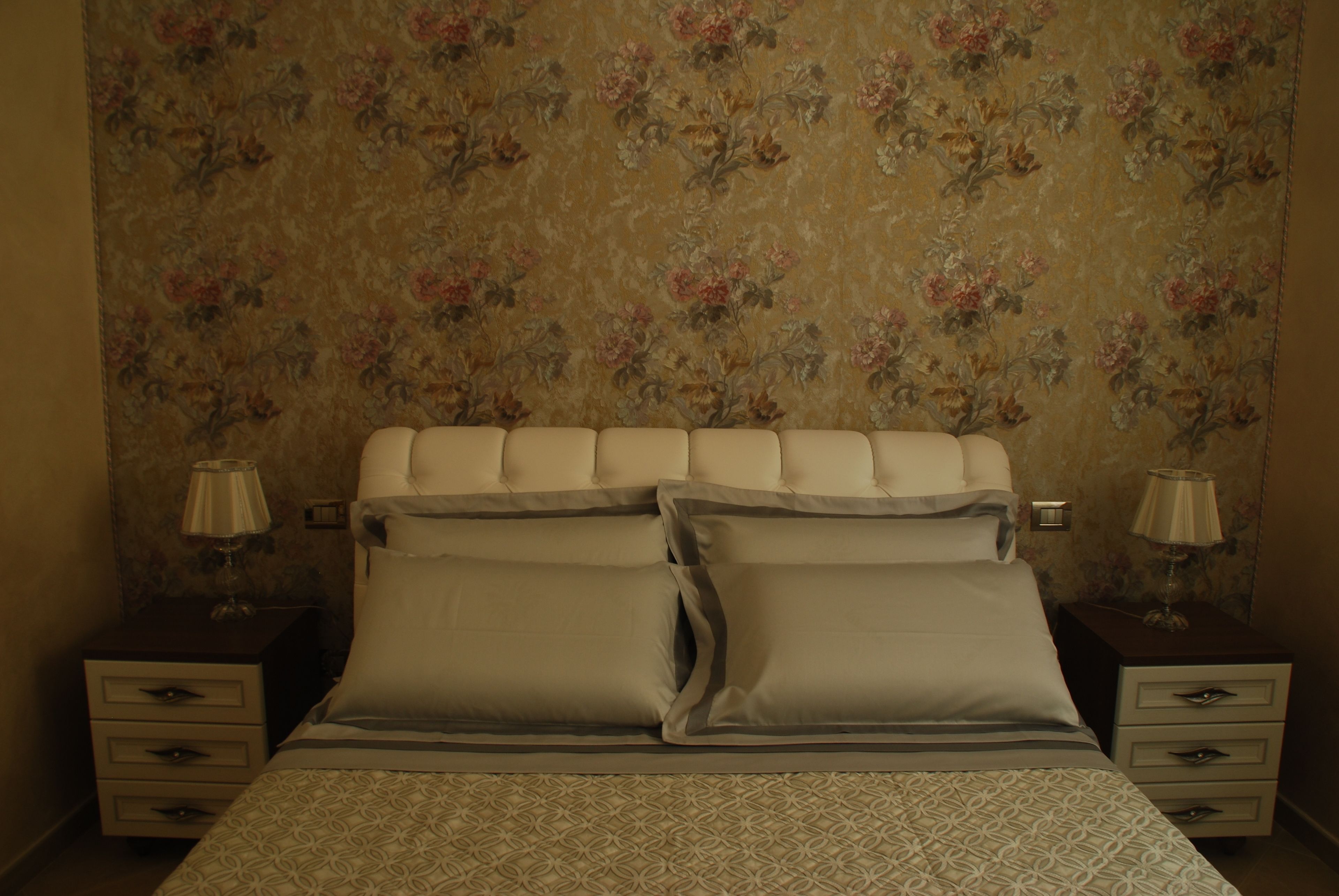 A neatly made bed with a patterned bedspread, two bedside tables with lamps, and a floral wallpaper backdrop.