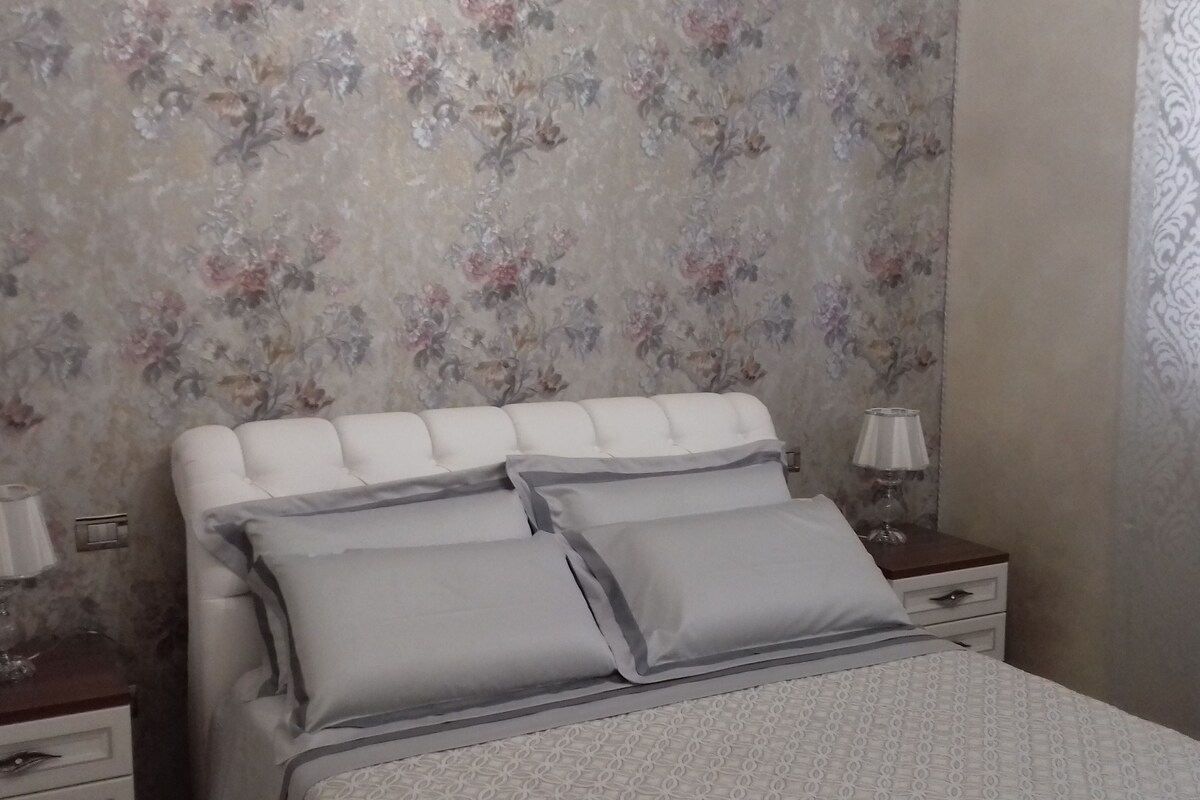 A bedroom with a floral wallpaper, a white upholstered headboard, two bedside tables with lamps, and a bed with grey bedding.