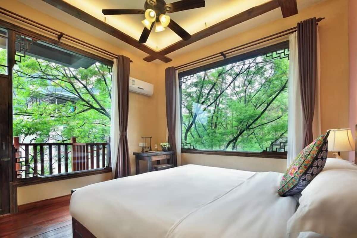 Une chambre à coucher avec un lit, deux fenêtres avec des rideaux, un ventilateur de plafond et une petite table avec une lampe.