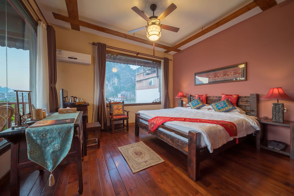 Une chambre avec un lit en bois, un ventilateur de plafond, une table recouverte d’une nappe, une chaise et une vue sur l’extérieur.
