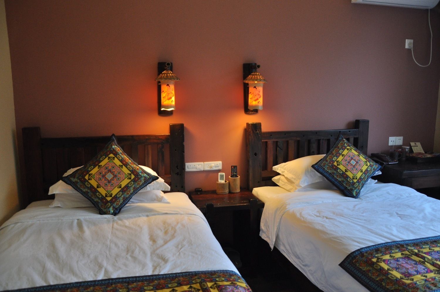 Quarto de hotel com duas camas, cada uma com almofada estampada, mesa de cabeceira com abajur e luminária na parede.