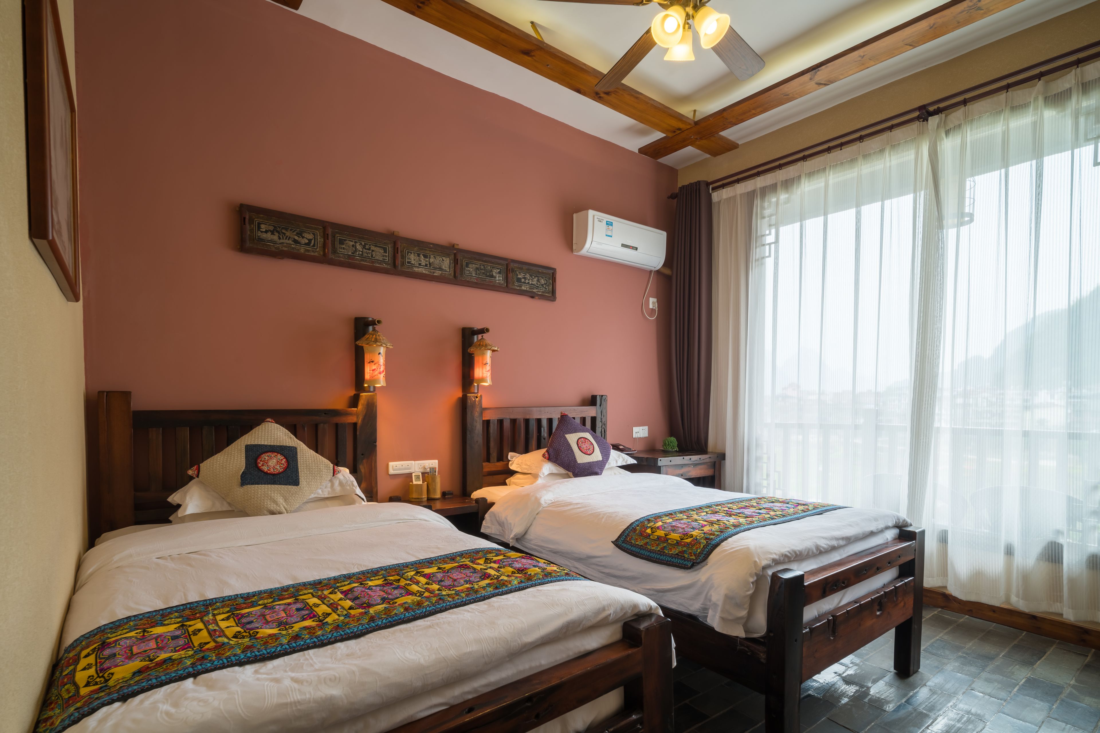 A room with two beds, a wooden ceiling, a wall-mounted air conditioner, and a window with sheer curtains.