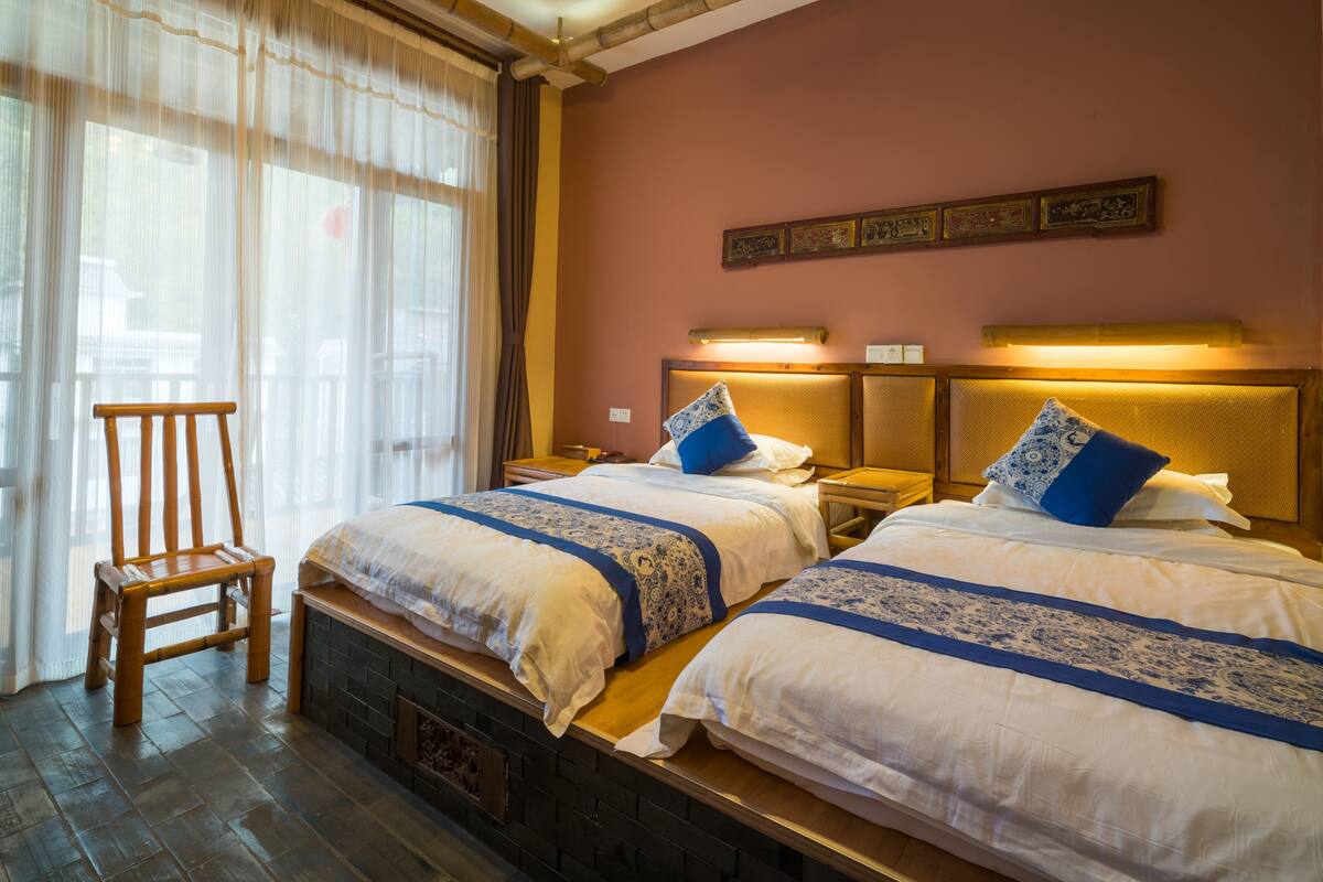 Une chambre d’hôtel avec deux lits, une chaise en bois et une fenêtre avec des rideaux légers.