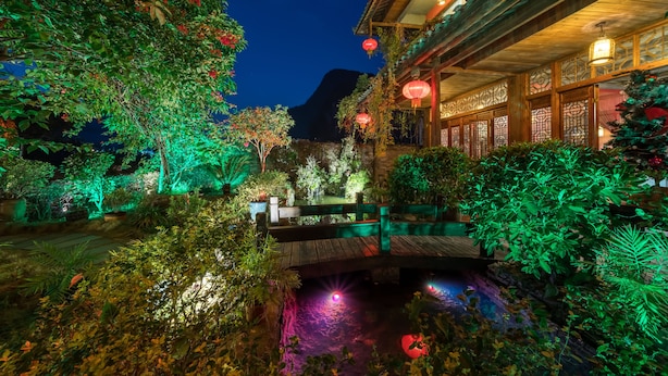 Ein traditioneller Innenhof mit einer Holzbrücke über einem Wasserspiel, umgeben von üppiger Vegetation und beleuchtet durch rote Laternen.