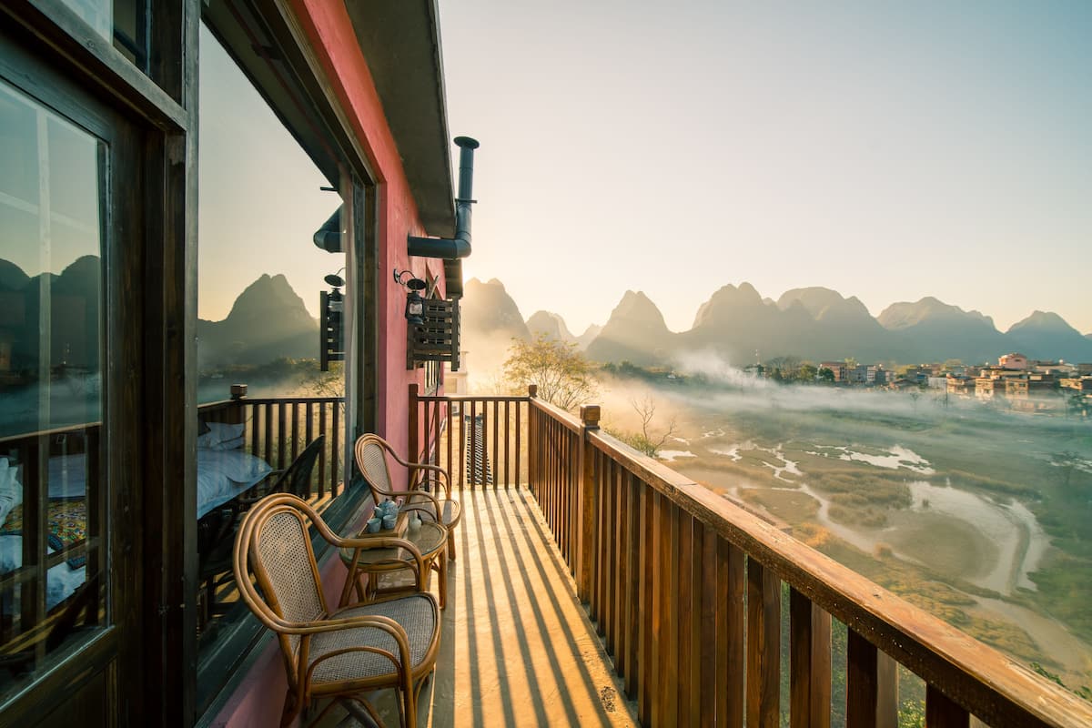 Un balcon doté de chaises en osier et d’une balustrade en bois, offrant une vue sur une rivière et des montagnes.