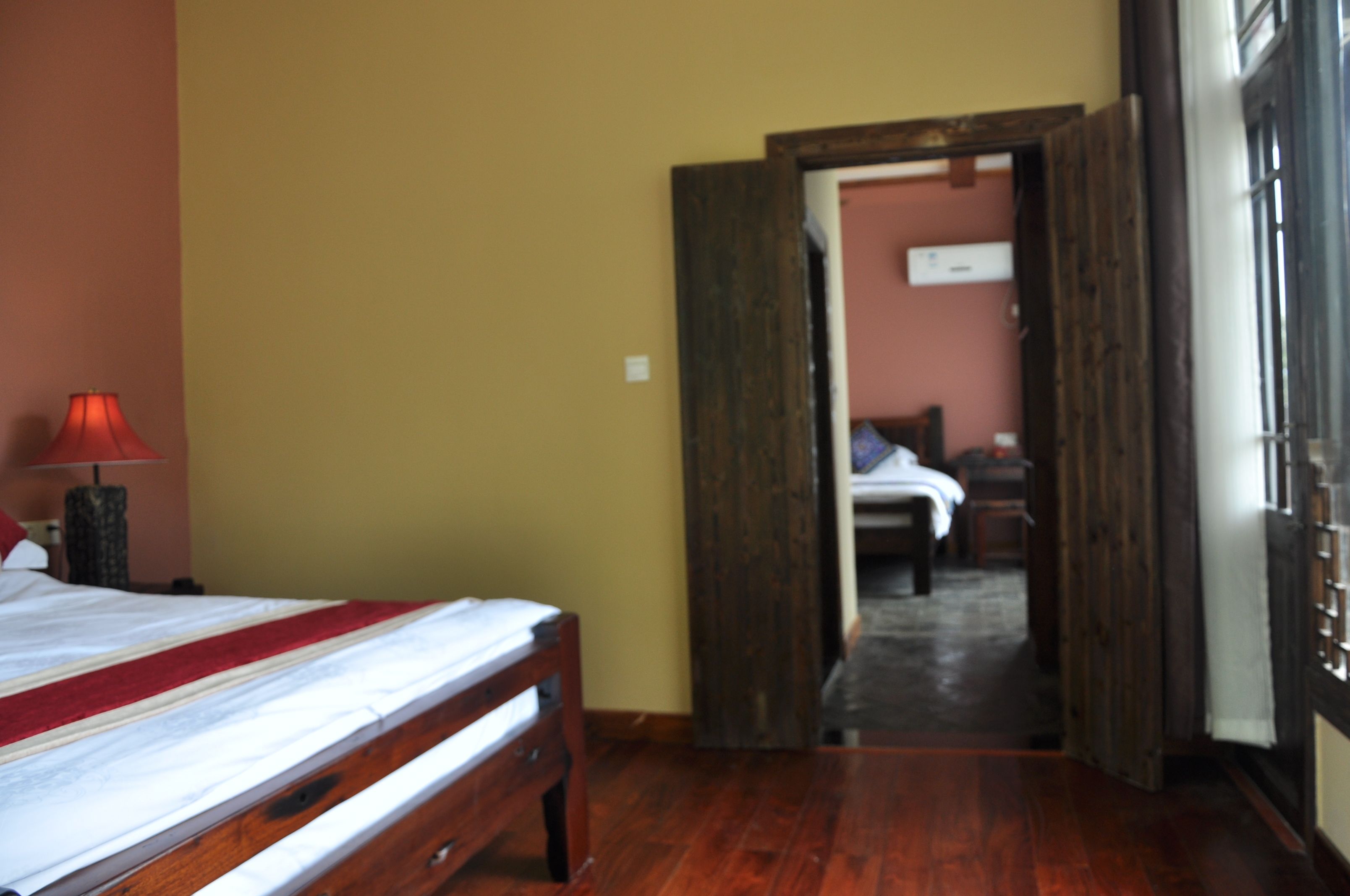 Una habitación de hotel con una cama de madera, una lámpara roja y vistas a otra habitación por una puerta abierta.