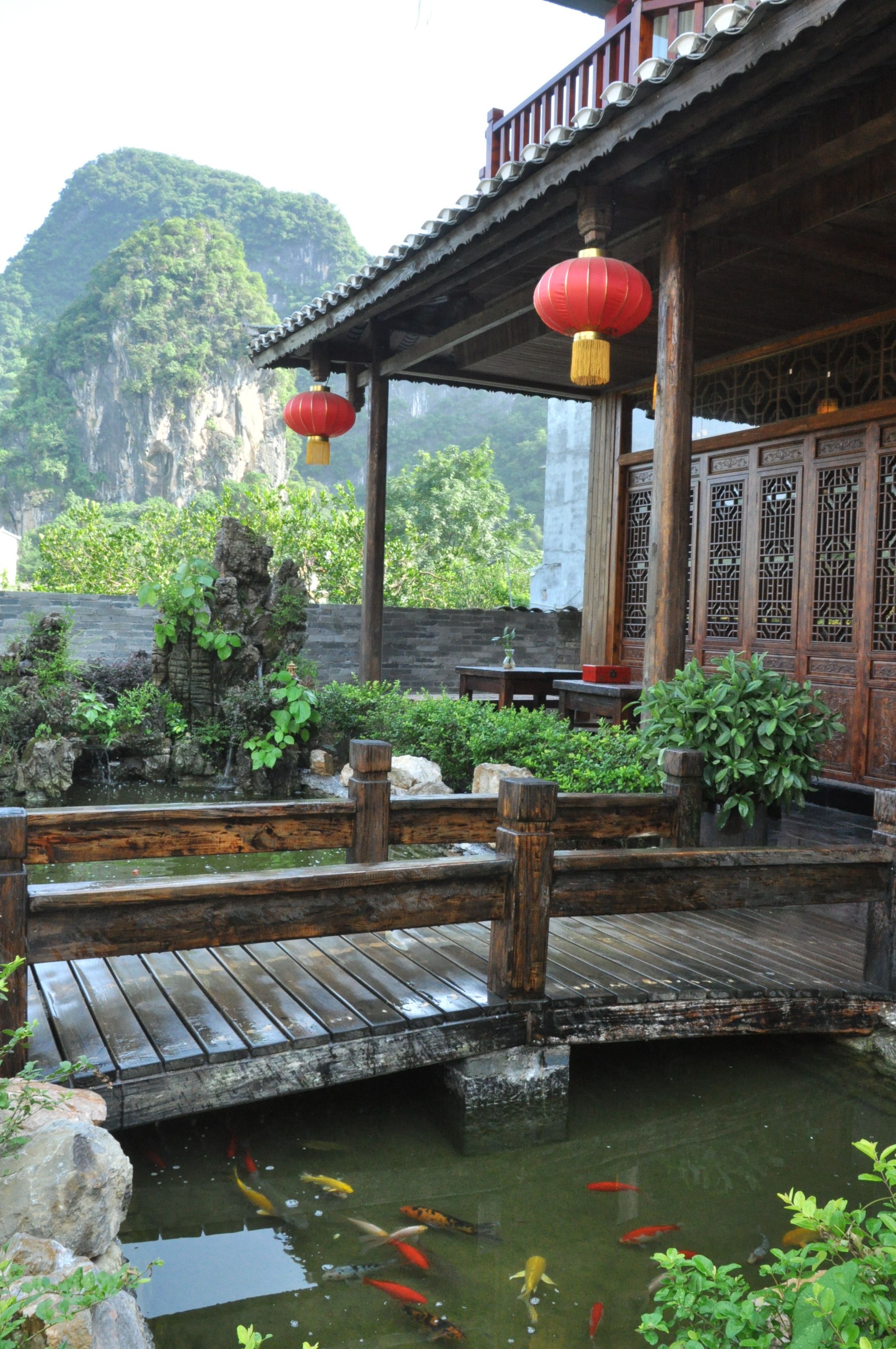 A traditional wooden pavilion with a koi pond, a wooden bridge, and a mountainous backdrop.